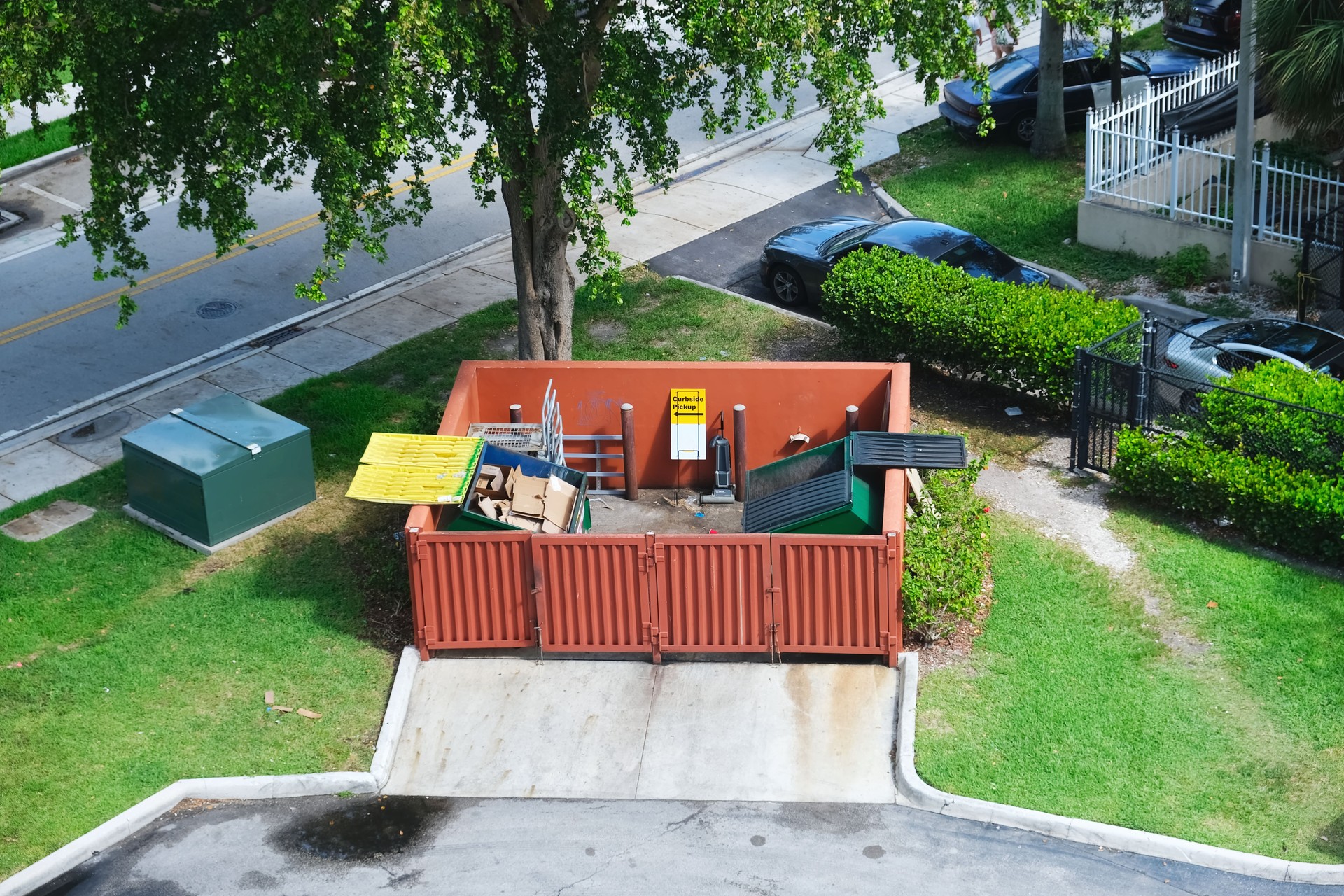 Dumpster half-filled with garbage next to residential area, road and parking lot behind commercial building aerial street view photo. Waste management concept.